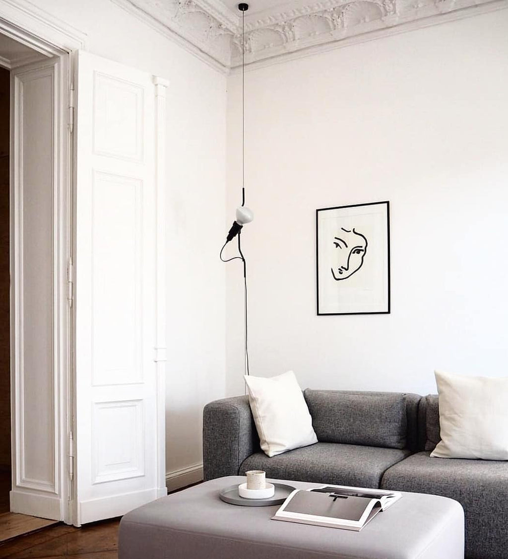 A Clérachic pendant spotlight hanging in a white room with a high moulded ceiling, above a grey sofa and a line drawing in a frame.