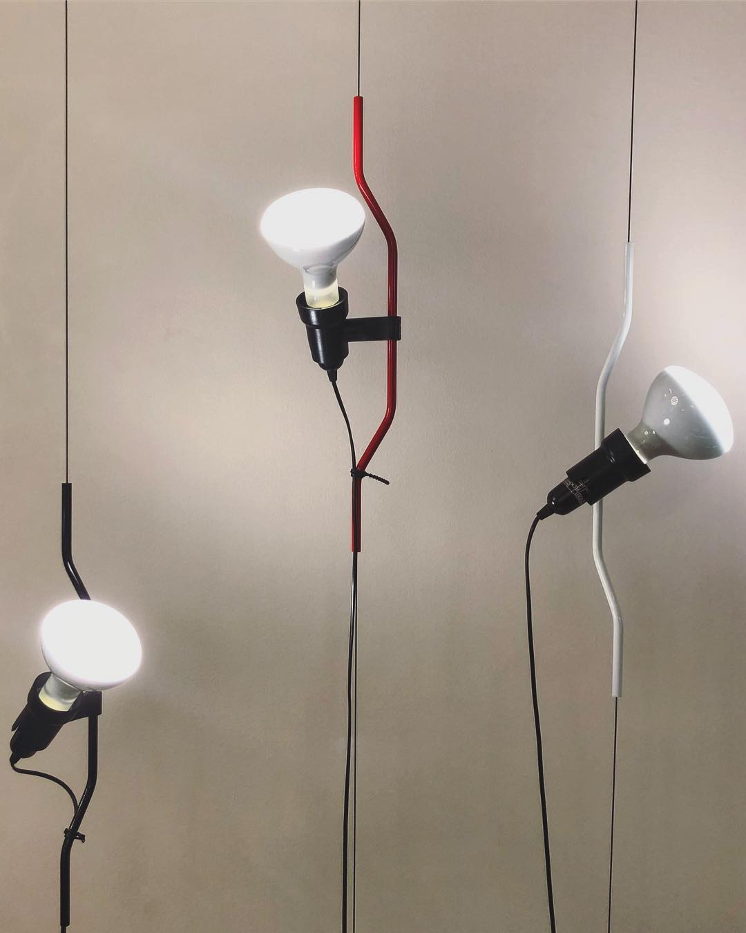 Three Clérachic pendant spotlights with white shades, one on a red cord and others in black and white, against a warm grey wall.