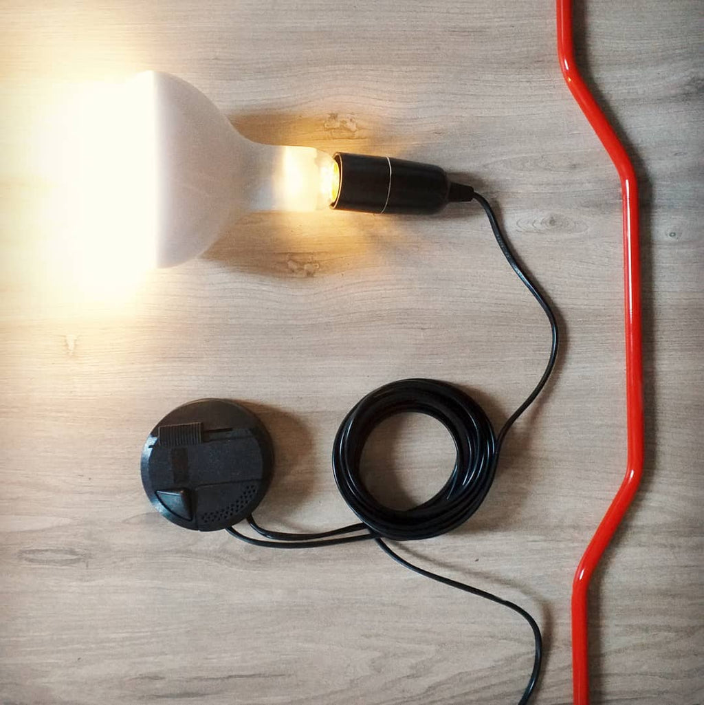 Top view of a Clérachic pendant with a red cord, a lit white bulb in a black socket and a coiled black lead, on a wooden floor.