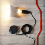 Top view of a Clérachic pendant with a red cord, a lit white bulb in a black socket and a coiled black lead, on a wooden floor.