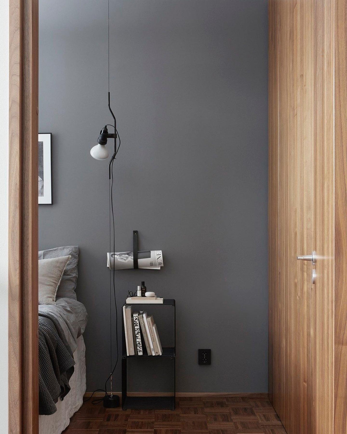 Lit Clérachic pendant with a white bulb hanging in a bedroom corner beside a slate-grey wall, above a slim black side table.