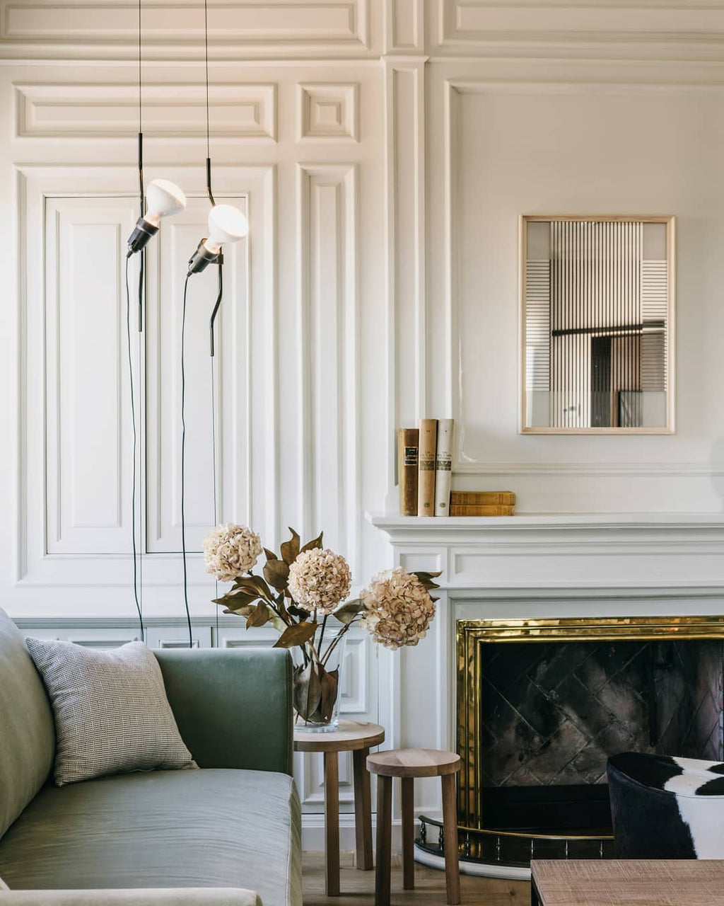Two Clérachic pendant spotlights with white shades hanging in front of a panelled white wall, beside a fireplace and a green sofa.