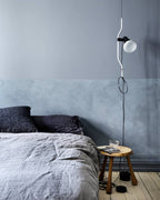 Clérachic pendant with a white bendable cord and a white bulb in a bedroom with a grey-blue wall, above a linen bed and a wooden stool.
