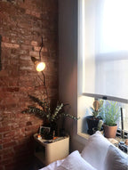 Lit Clérachic pendant with a warm bulb beside a red brick wall and a window, above a bedside shelf with plants.