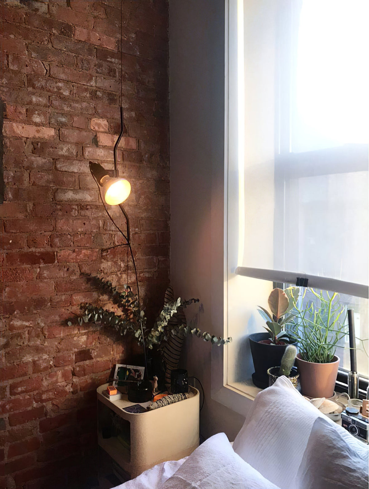 Lit Clérachic pendant with a warm bulb beside a red brick wall and a window, above a bedside shelf with plants.