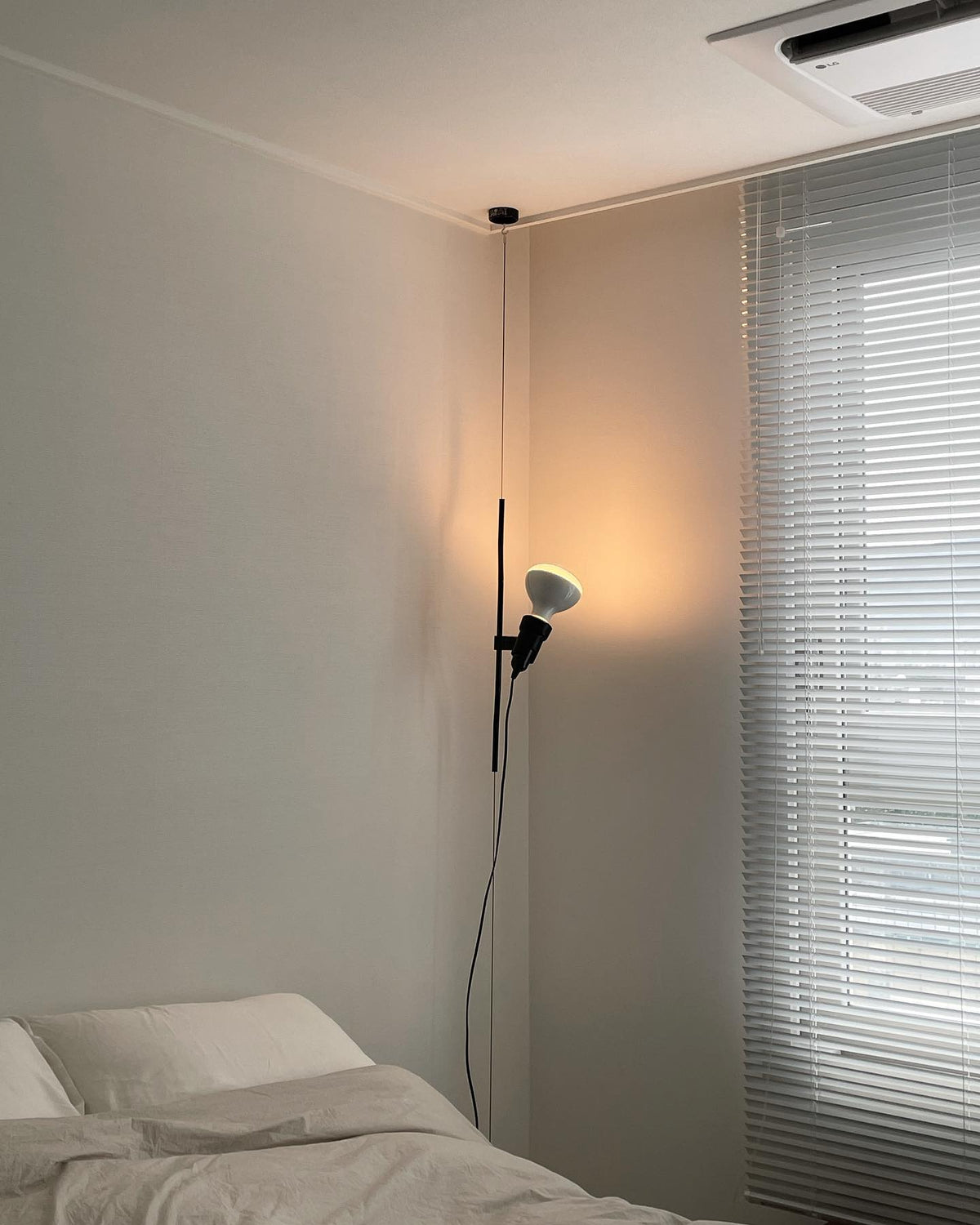 A Clérachic pendant spotlight lit in a bedroom corner, with a venetian blind at the window and a ceiling air unit above.