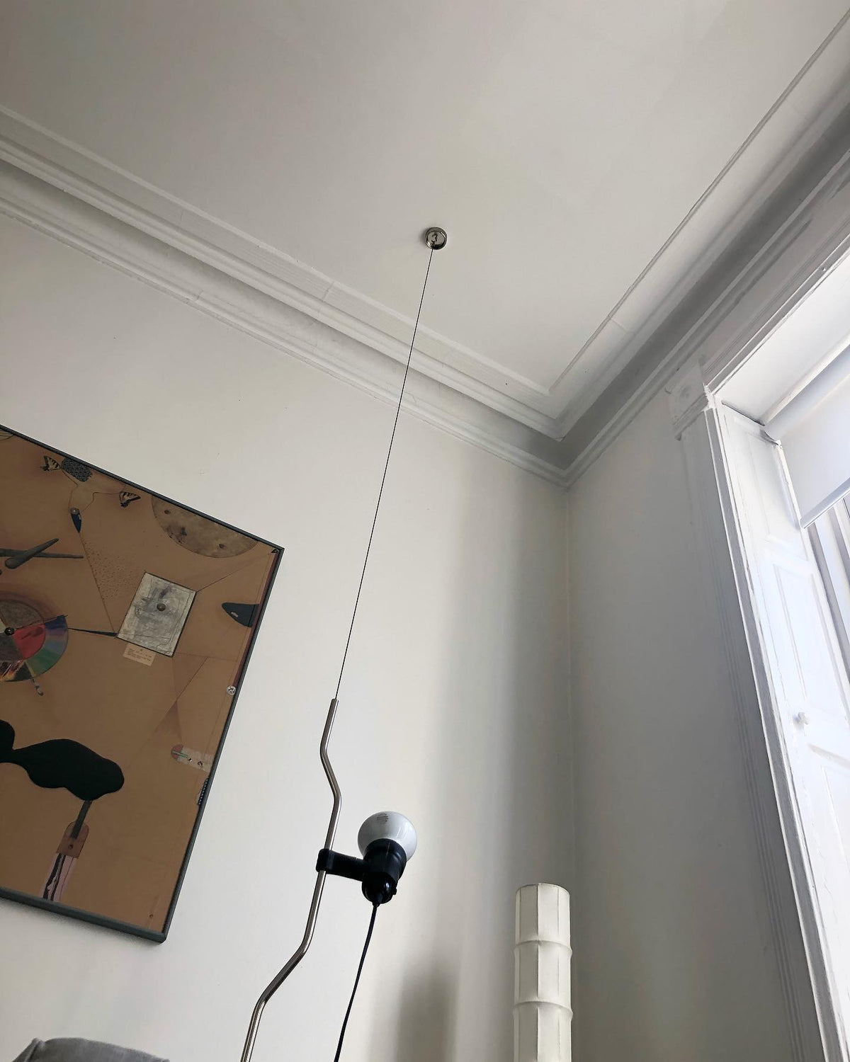 Clérachic pendant with a bendable cord and a white bulb, seen from below against a ceiling with plaster mouldings and a framed picture.