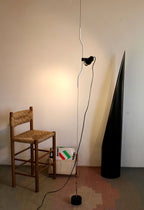 A Clérachic pendant with a white bendable cord section and a black spotlight head, falling to a base beside a wooden rush-seat chair.