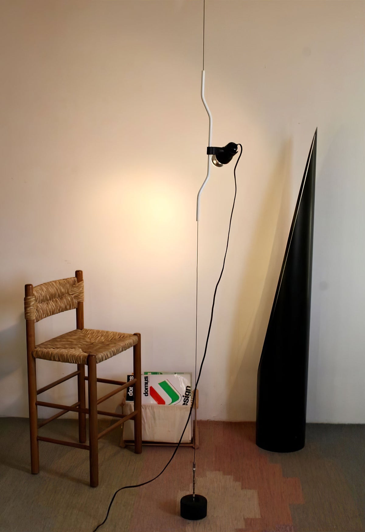 A Clérachic pendant with a white bendable cord section and a black spotlight head, falling to a base beside a wooden rush-seat chair.