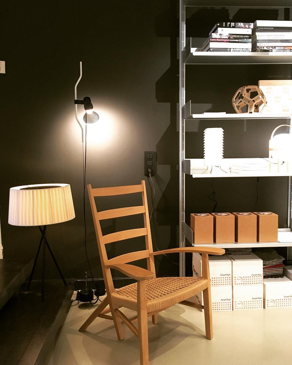 Lit Clérachic pendant spotlight on a cable beside a wooden armchair, a shelving unit and a fabric-shaded floor lamp in a dark room.