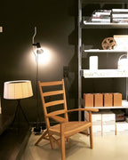 Lit Clérachic pendant spotlight on a cable beside a wooden armchair, a shelving unit and a fabric-shaded floor lamp in a dark room.