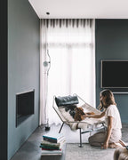 Clérachic pendant with a bendable cord hanging by sheer curtains in a slate-green living room, with a lounge chair and a seated woman and dog.