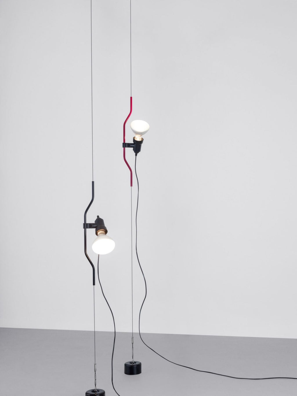 Two Clérachic pendants, one with a red bendable cord section and one in black, hanging side by side on cables against a plain wall.