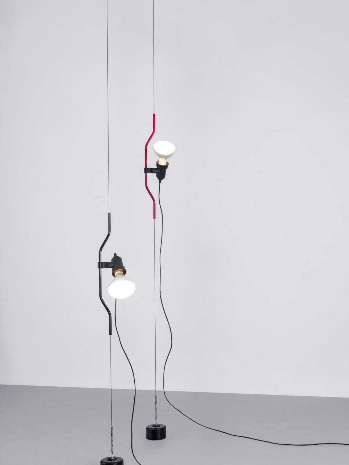 Two Clérachic pendants, one with a red bendable cord section and one in black, hanging side by side on cables against a plain wall.