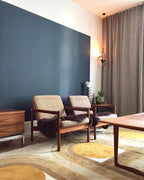 A Clérachic pendant spotlight lit beside heavy grey curtains, above wooden armchairs, a low table and a patterned rug in a room with a blue wall.