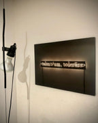 Close view of a Clérachic pendant with a black bendable cord and a white bulb, beside a dark wall sign with softly lit text.