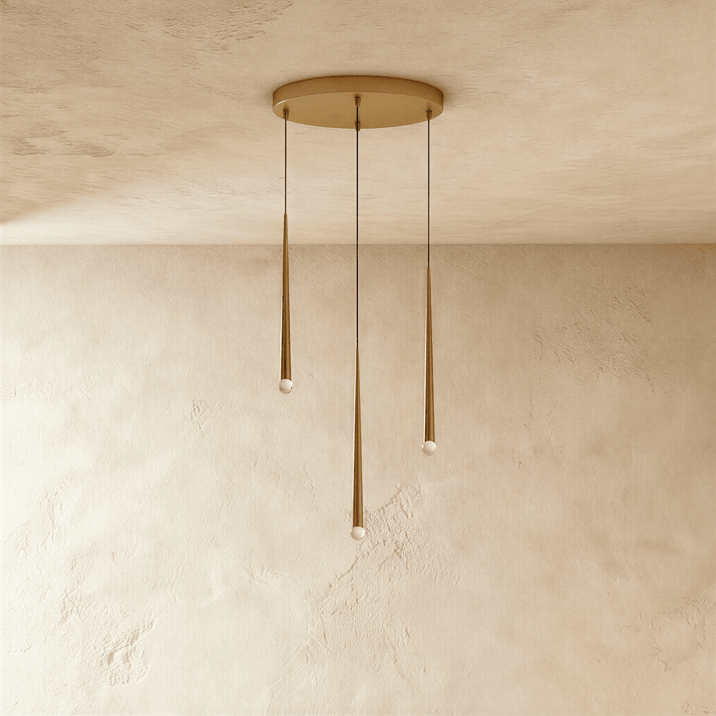 Clérachic three-light gold cone pendant cluster on a round brass-toned canopy, tips at staggered heights against pale limewashed plaster.