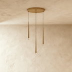 Clérachic three-light gold cone pendant cluster on a round brass-toned canopy, tips at staggered heights against pale limewashed plaster.