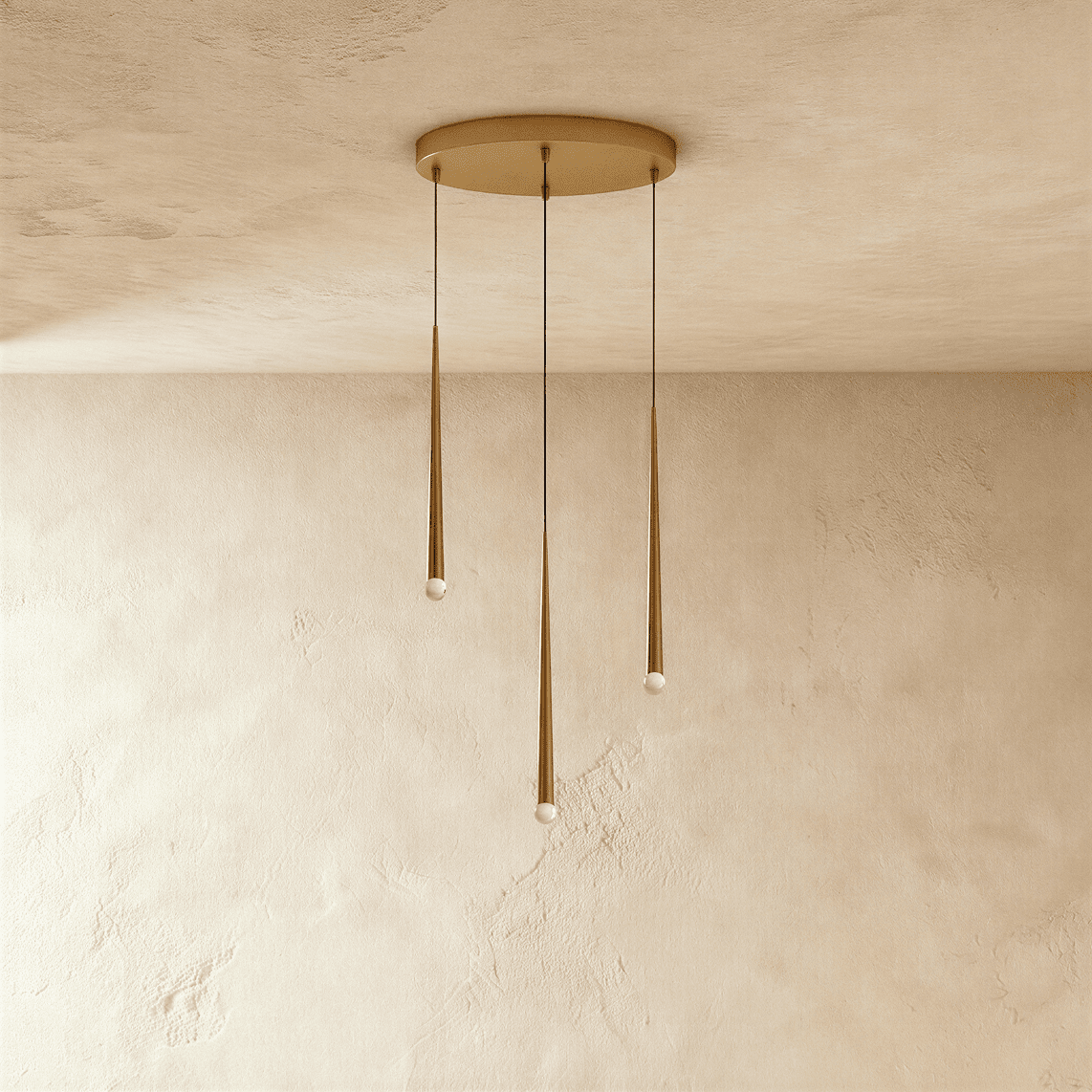 Clérachic three-light gold cone pendant cluster on a round brass-toned canopy, tips at staggered heights against pale limewashed plaster.