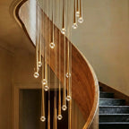 Close view of Clérachic gold cone pendants with faceted crystal tips at varied heights beside a curved walnut stair rail.