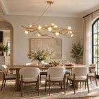 A Clérachic branch chandelier glowing above a long wooden dining table with cream chairs, in a room with an arched window and framed art.