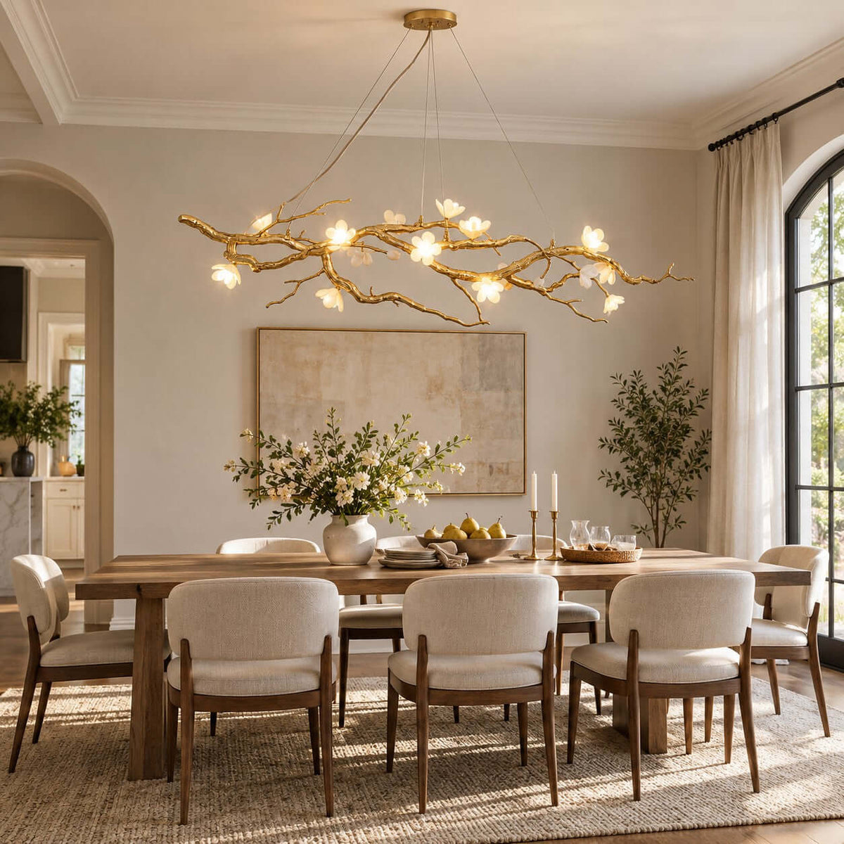 A Clérachic branch chandelier glowing above a long wooden dining table with cream chairs, in a room with an arched window and framed art.