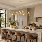 A Clérachic gold branch chandelier hangs above an island with leather-seat stools, in a kitchen with tall windows and a flowering branch.