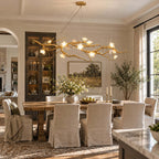 Dining room with a Clérachic gold branch chandelier over a rustic table with slipcovered chairs, a glass display cabinet and a landscape painting.