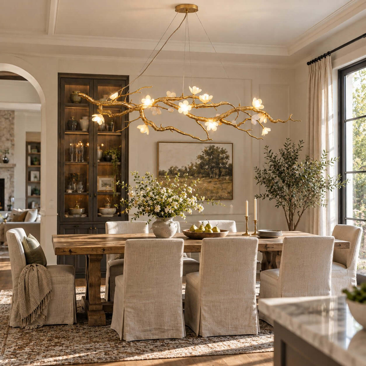 Dining room with a Clérachic gold branch chandelier over a rustic table with slipcovered chairs, a glass display cabinet and a landscape painting.
