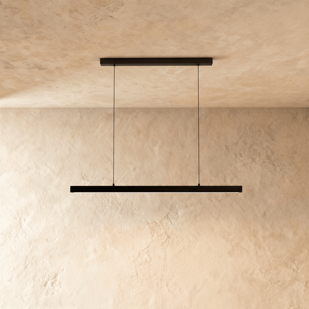 Clérachic black linear pendant, cable-suspended, centred against a textured limewash wall in soft daylight.