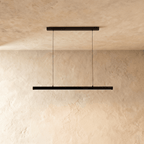Clérachic black linear pendant, cable-suspended, centred against a textured limewash wall in soft daylight.