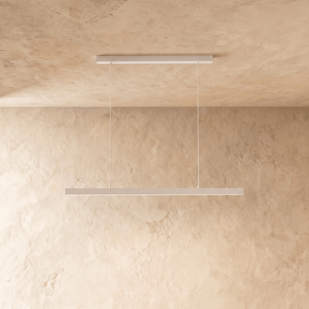 Clérachic white linear pendant, cable-suspended, centred against a textured limewash wall in soft light.