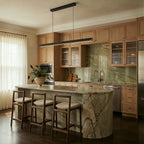 Clérachic black linear pendant hangs above a marble kitchen island with oak cabinetry and a green stone backsplash.