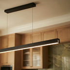 Clérachic black linear pendant glows warm above oak kitchen cabinets, its bar lit beside a stone backsplash.