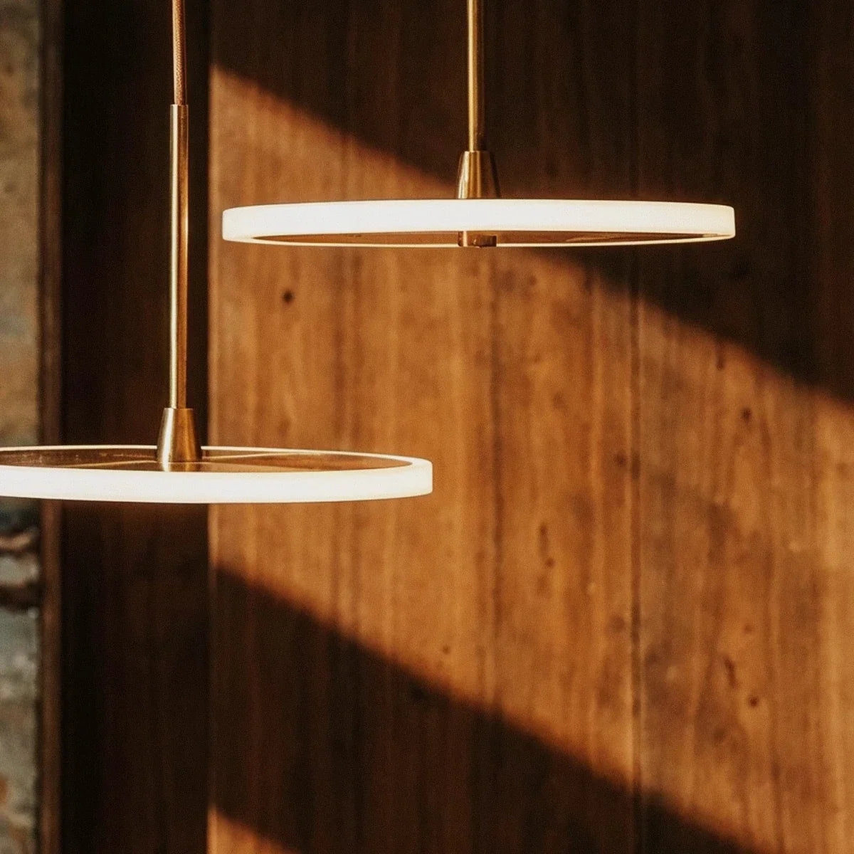 Clérachic close-up of two gold LED discs with frosted edges on brass stems, lit against sunlit wooden panelling.