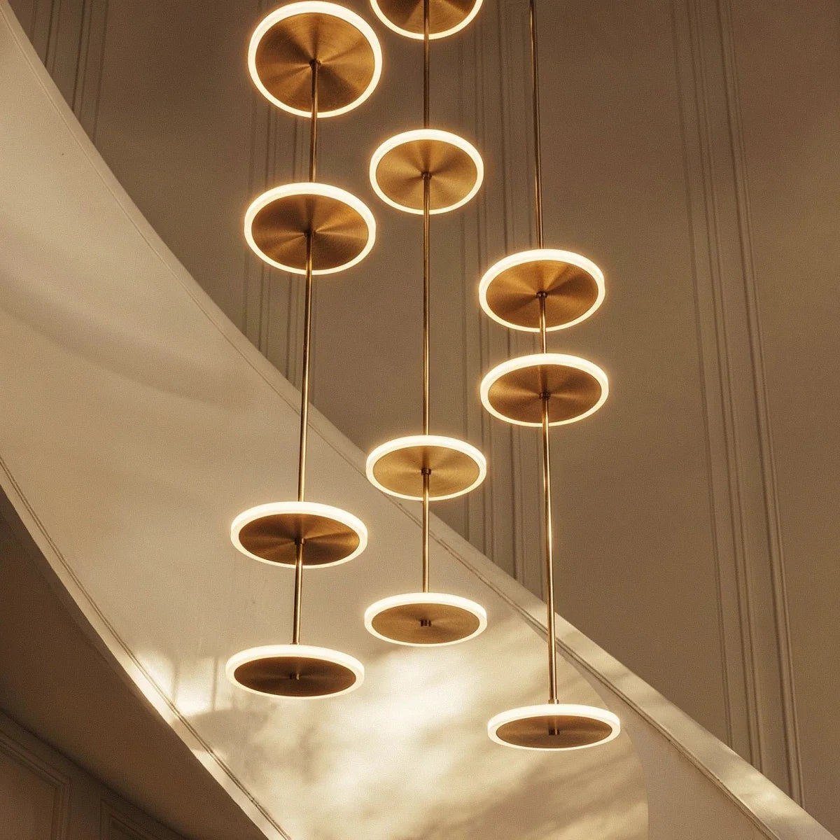 Clérachic underside view of glowing gold LED discs with brushed centres, strung on thin rods beside a curved staircase.