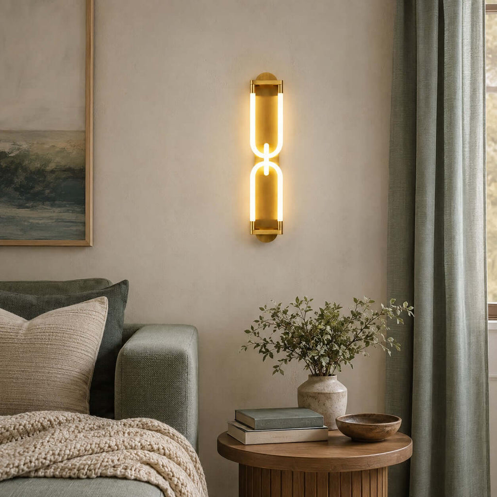 A Clérachic gold wall light with two linked glowing loops, hung beside a sage curtain above a round wooden side table.