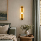 A Clérachic gold wall light with two linked glowing loops, hung beside a sage curtain above a round wooden side table.