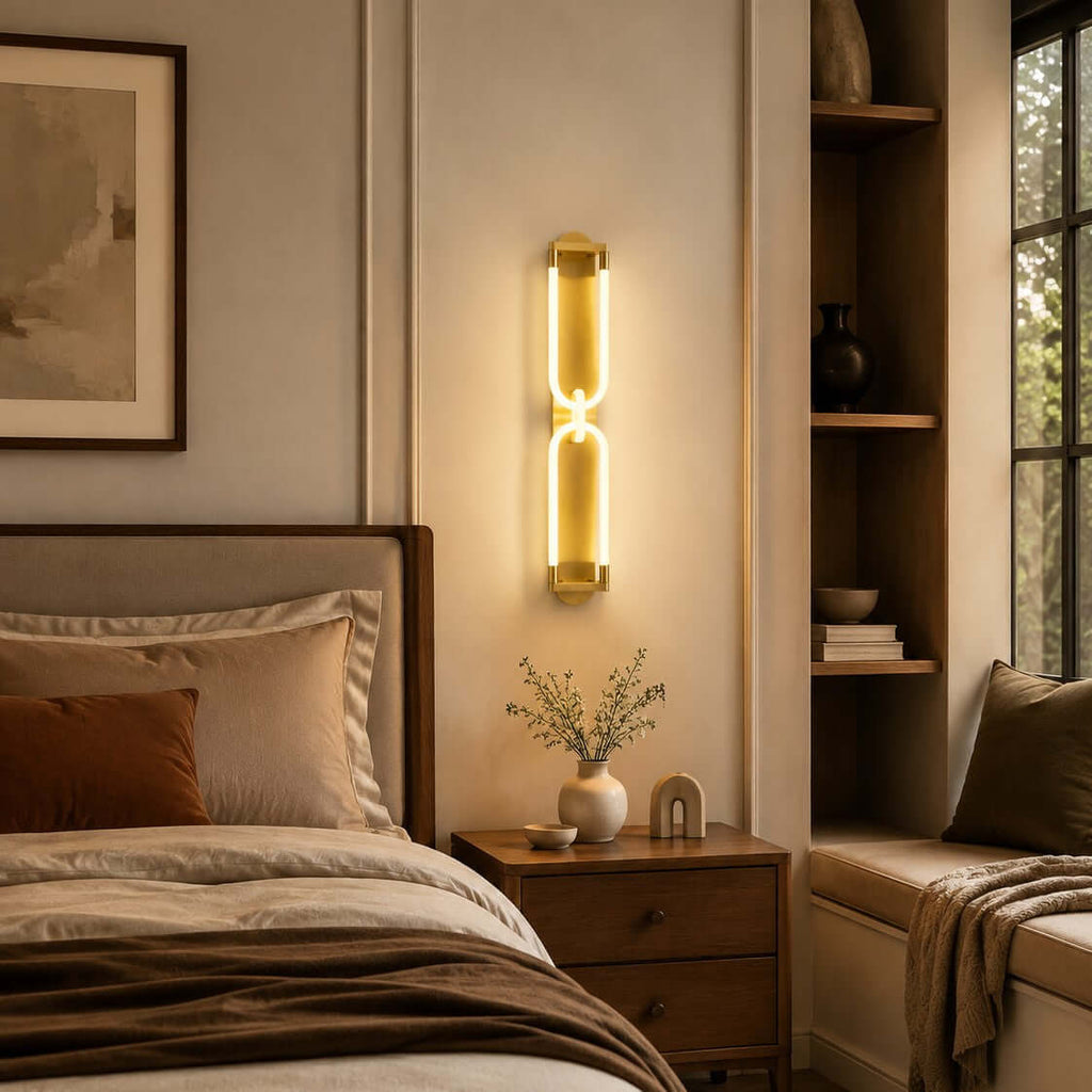 Lit gold Clérachic loop wall light beside a bed, over a small vase and a nightstand, with a built-in shelf at the right.