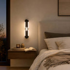 Black Clérachic loop wall light glowing beside a cream headboard, above a floating wooden nightstand with an open book.