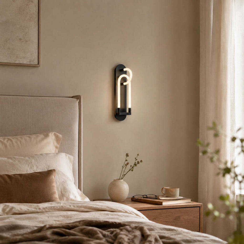 A black Clérachic wall light lit above a cream-headboard bed, with a vase and a cup on the wooden nightstand below.