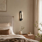 A black Clérachic wall light lit above a cream-headboard bed, with a vase and a cup on the wooden nightstand below.