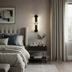 A lit black Clérachic wall light above a bedside table, next to a grey tufted headboard and long cream curtains.