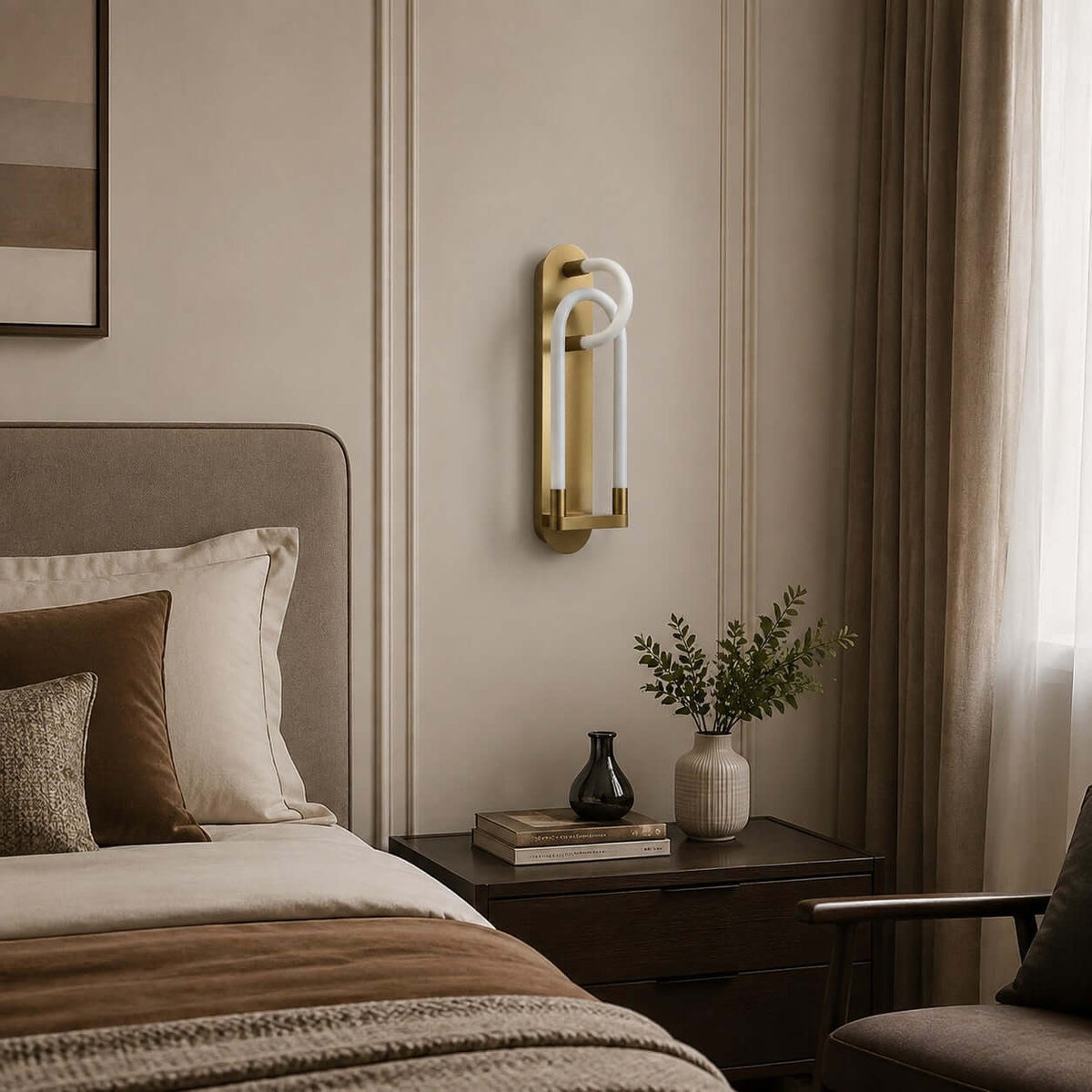 Gold Clérachic loop wall light, unlit, on a cream panelled wall beside a bed, with a vase and books on a dark table.