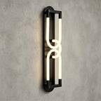 Close view of a lit black Clérachic wall light, two linked frosted loops on a slim frame against grey plaster.
