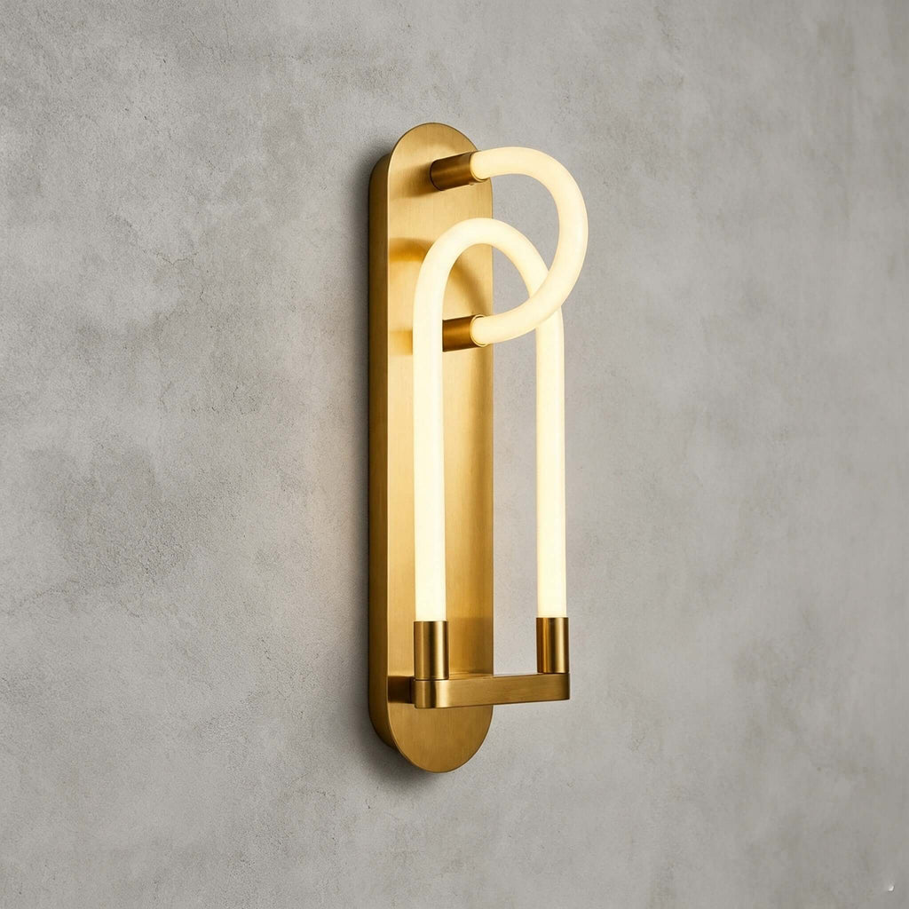 A gold Clérachic loop wall light seen from an angle, the frosted tube glowing warm in front of a brushed brass backplate.