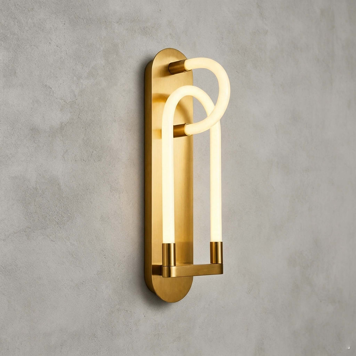 A gold Clérachic loop wall light seen from an angle, the frosted tube glowing warm in front of a brushed brass backplate.