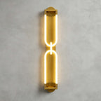 Tall gold Clérachic wall light lit on grey plaster, two stacked loops joined by a small link, with a warm halo around the frame.