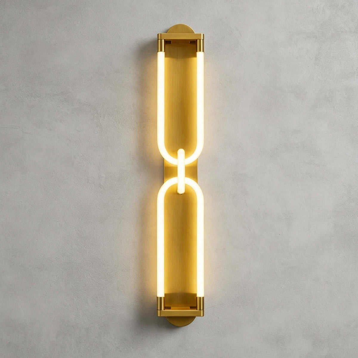 Tall gold Clérachic wall light lit on grey plaster, two stacked loops joined by a small link, with a warm halo around the frame.