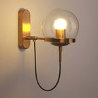 This wall-mounted Clérachic sconce in brushed bronze glows warm white against the wall, its looped black cable visible below the globe.